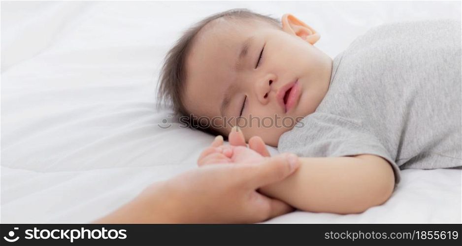 Young asian mother touch hand of little baby girl with tender on bed in the bedroom, mom love newborn and care, woman with expression with child together, parent and daughter, family concept.