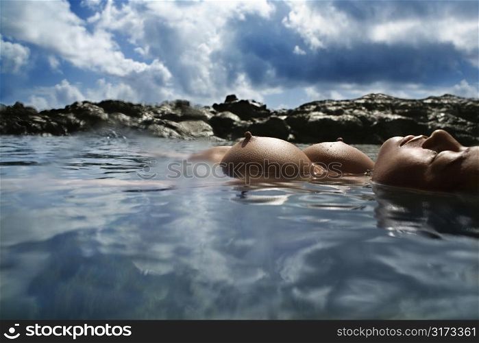 Young Asian nude woman floating on back on rocky coast.