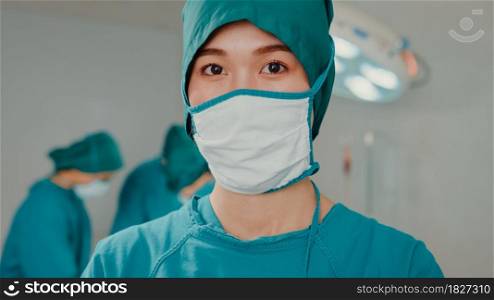 Young Asian nurse woman looking at camera and smiling after performing invasive surgery on patient in hospital operating room. Diverse team of professional surgeon, Healthcare and medical concept.
