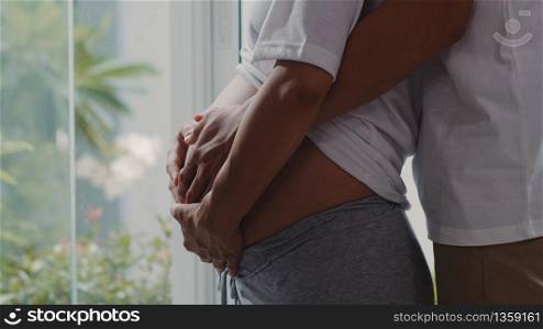 Young Asian Pregnant couple hug and holding belly talking with their child. Mom and Dad feeling happy smiling peaceful while take care baby, pregnancy near window in living room at home concept.