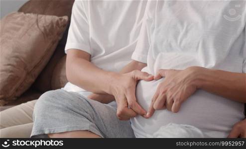 Young Asian Pregnant couple making heart sign holding belly. Mom and Dad feeling happy smiling peaceful while take care baby, pregnancy lying on sofa in living room at home concept.
