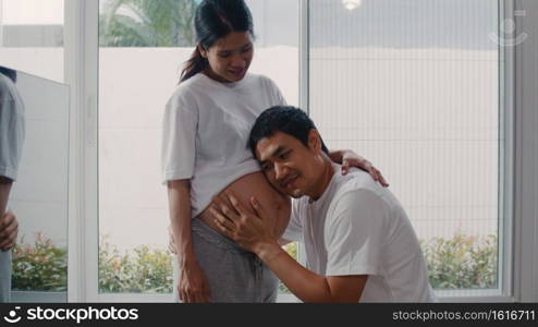 Young Asian Pregnant couple man kissing his wife belly talking with his child. Mom and Dad feeling happy smiling peaceful while take care baby, pregnancy near window in living room at home concept.