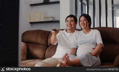 Young Asian Pregnant couple watching TV. Mom and Dad feeling happy smiling peaceful while take care baby, pregnancy lying on sofa in living room at home concept.