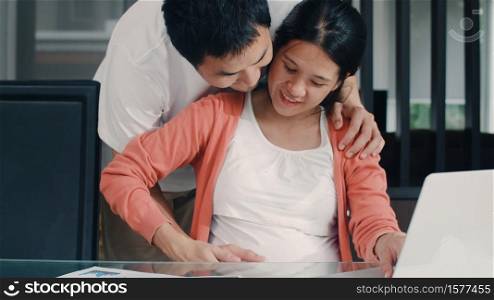 Young Asian Pregnant woman using laptop records of income and expenses at home. Dad touch his wife belly while record budget, tax, financial document working in living room at home concept.