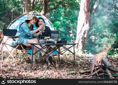 Young Asian pretty woman and her girlfriend sitting at front of tent, use mobile phone take photo during camping in forest with happiness together