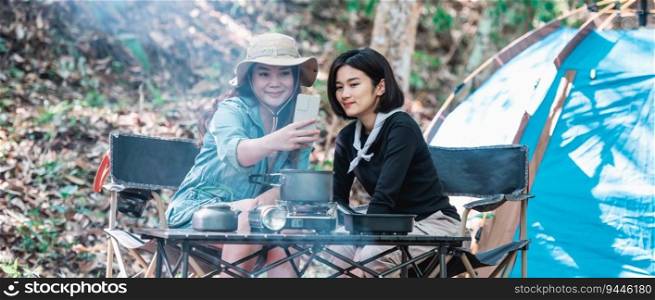 Young Asian pretty woman and her girlfriend sitting at front of tent, use mobile phone take photo during camping in forest with happiness together
