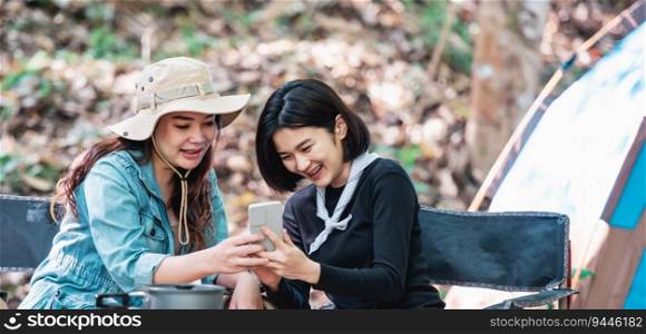 Young Asian pretty woman and her girlfriend sitting at front of tent, use mobile phone take photo during c&ing in forest with happiness together
