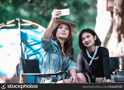 Young Asian pretty woman and her girlfriend sitting at front of tent, use mobile phone take photo during c&ing in forest with happiness together