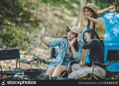 Young Asian pretty woman and her girlfriend sitting at front of tent, use mobile phone take photo during c&ing in forest with happiness together