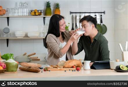 Young Asian romantic couple is cooking in the kitchen. Happy beautiful woman holding milk of glass and feeding milk to her handsome man while making healthy breakfast food at home together