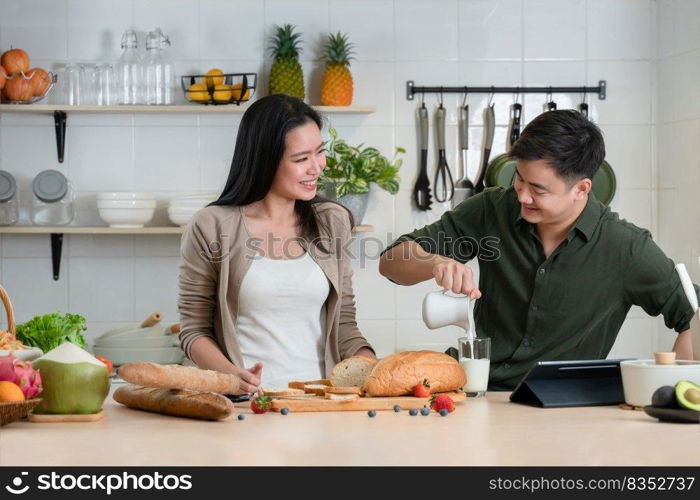 Young Asian romantic couple is cooking in the kitchen. A beautiful woman and handsome man making healthy breakfast food while boyfriend pouring milk into glass for his girlfriend