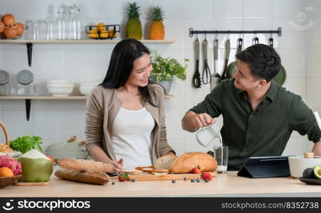 Young Asian romantic couple is cooking in the kitchen. A beautiful woman and handsome man making healthy breakfast food while boyfriend pouring milk into glass and smile at his girlfriend