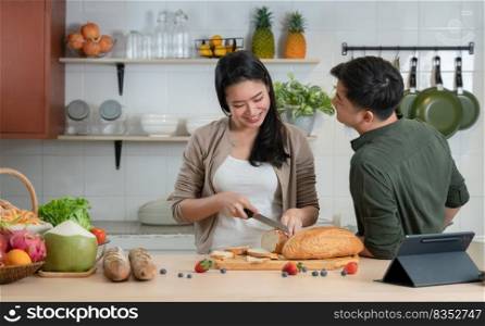 Young Asian romantic couple is cooking in the kitchen. Happy beautiful woman making healthy food like bread, fruits and salad for her handsome man while boyfriend looking at girlfriend with love