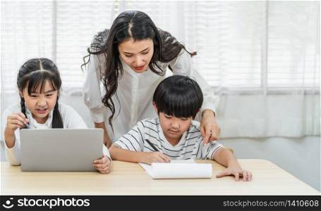 Young Asian Single Mother teaching or helping daughter and son doing home work with technology laptop in home, home school education, relationship and family, communication and conversation concept