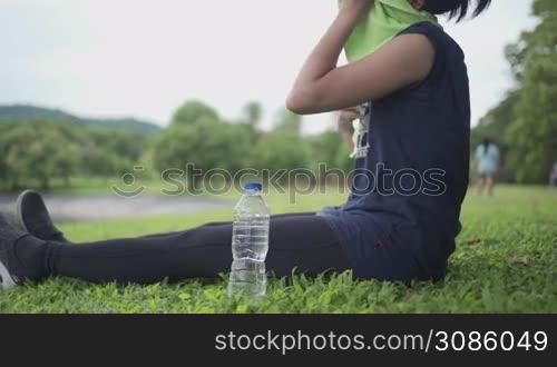 Young asian sportive woman resting sweat after workout, sit down on comfortable grass, sipping mineral water and enjoying green fresh view of public park, health care routine, active lifestyle concept