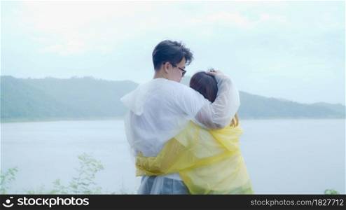 Young Asian sweet couple feeling happy using romantic time playing rain while wearing raincoat standing near lake. Lifestyle couple enjoy and relax in rainy day.