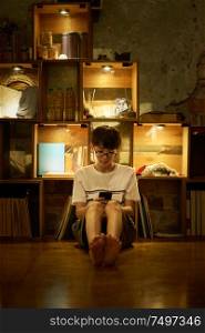 Young asian teenage boy using smartphone sit in front of bookshelf cabinet with warm lighting