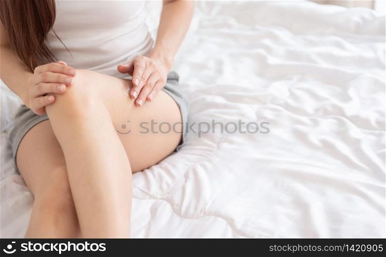 Young Asian woman applying smooth body lotion on her legs while sitting in bed at her bedroom. Copy space. Beauty, Body and Health care.
