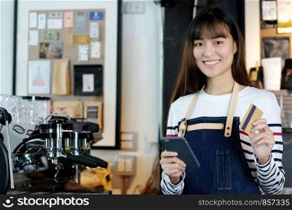 Young asian woman barista using smartphone and holding credit card at cafe counter, food and drink business concept
