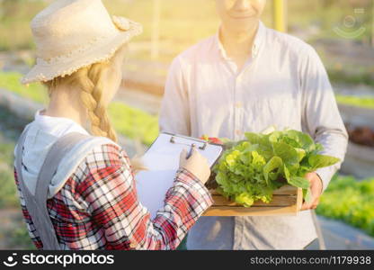 Young asian woman checking vegetable organic hydroponic farm and man harvest picking up fresh vegetable, girl writing record document grow of leaf for quality produce, small business owner concept.