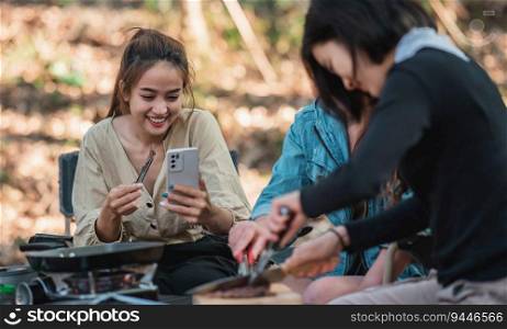 Young asian woman cooking and her friend enjoy to make the meal in pan, They are talk and laugh with fun together while camping in nature park