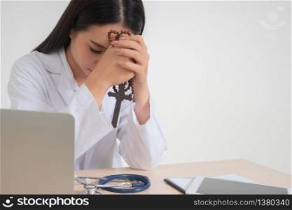Young Asian woman doctor sitting on desk and holding a cross and praying, she looks unhappy and tired because of overwork, Concept of stressed exhausted and doctor liability, Dealing with frustrated