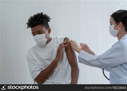 Young Asian woman doctor wear mask is giving injections or vaccines to arm of African American boy who fear syringe and turn face away at hospital. Preventing spread of COVID-19 by vaccinating people