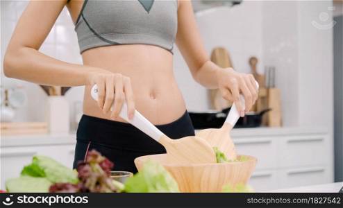 Young Asian woman drink orange juice making salad in the kitchen, beautiful female in sport clothing use organic fruits and vegetables making healthy food by herself at home. Healthy food concept.