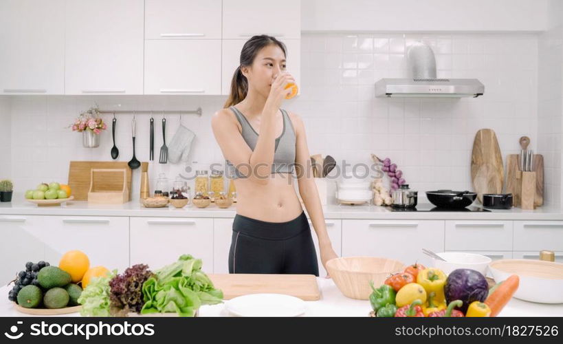 Young Asian woman drinking orange juice in the kitchen, beautiful female in sport clothing use organic fruits lots of nutrition making orange juice by herself at home. Healthy food concept.