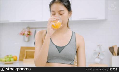 Young Asian woman drinking orange juice in the kitchen, beautiful female in sport clothing use organic fruits lots of nutrition making orange juice by herself at home. Healthy food concept.
