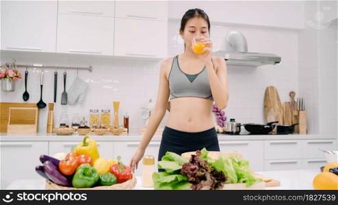 Young Asian woman drinking orange juice in the kitchen, beautiful female in sport clothing use organic fruits lots of nutrition making orange juice by herself at home. Healthy food concept.