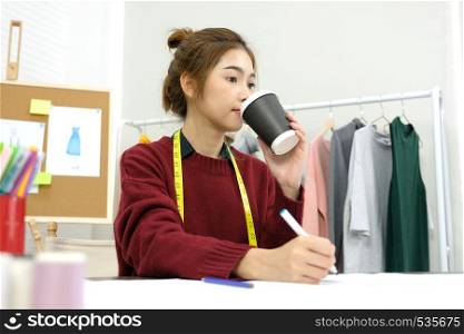 Young asian woman fashion designer working and drinking coffee in studio with happiness emotion, small business owner, fashion industry concept