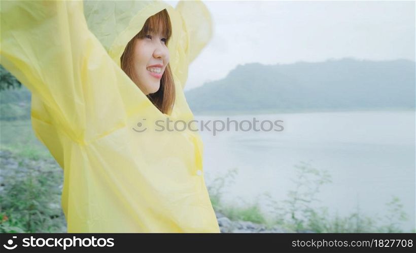 Young Asian woman feeling happy playing rain while wearing raincoat standing near lake. Lifestyle women enjoy and relax in rainy day.