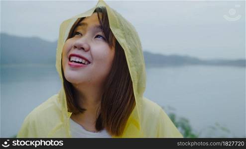 Young Asian woman feeling happy playing rain while wearing raincoat standing near lake. Lifestyle women enjoy and relax in rainy day.