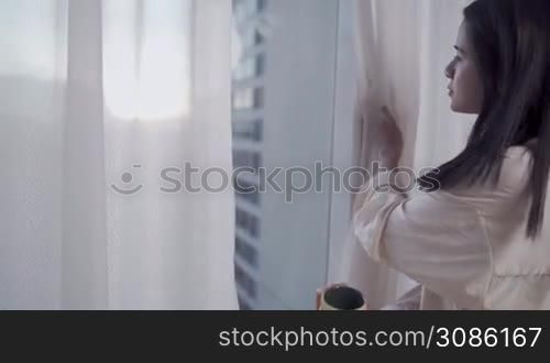 Young Asian woman get up in the early morning open window curtain, sipping coffee cup, female enjoy a cup of coffee and look through the window On high rise condo, urban city view during lockdown