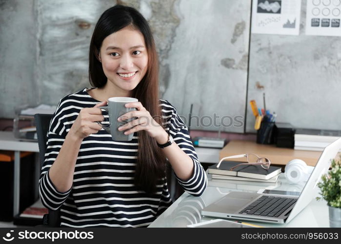 Young asian woman holding a coffee cup with smiling face, positive emotion at working desk background, casual office life, worink at home concept