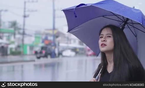 Young asian woman holding blue umbrella standing alone on the street side, waving hands calling or hitchhiking waiting for taxi or public transport service to pick up, girl with expectation