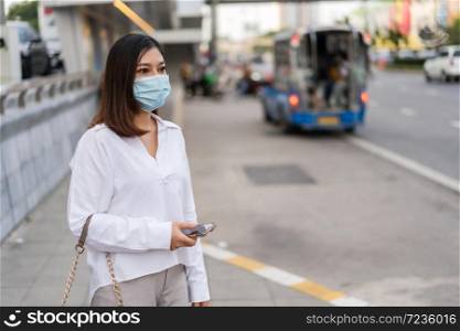 young asian woman holding smartphone and waiting for bus at bus stop in city street and wearing face mask protective for spreading of coronavirus(covid-19) pandemic, new normal concept