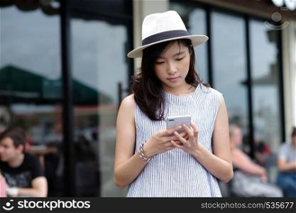 Young asian woman in casual style using smartphone while standing in front of cafe in the city outdoors background, people working outdoors with technology, urban lifestyle, people on phone