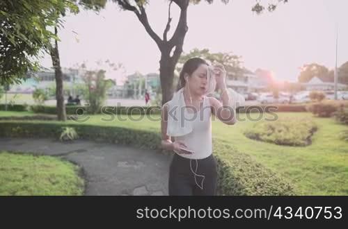 Young asian woman in sport clothing walking at the park during sunset golden hour, relaxing walk wiping sweat after hard work exercises, using phone singing to the music from earphones