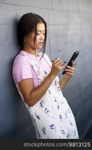 Young Asian woman in stylish clothes browsing news on mobile phone with stylus while leaning on gray wall on city street. Asian female leaning on wall and using cellphone