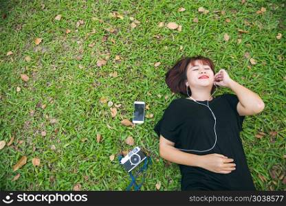 Young Asian woman laying on the green grass listening to music i. Young Asian woman laying on the green grass listening to music in the park with a chill emotion. Young woman relaxing on the grass with her music playlist. Outdoor activity in the park concept.