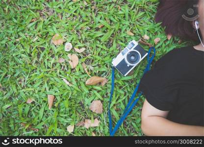 Young Asian woman laying on the green grass listening to music in the park with a chill emotion. Young woman relaxing on the grass with her camera beside. Outdoor activity in the park concept.