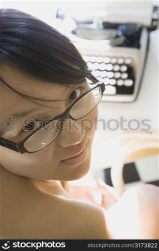 Young Asian woman making eye contact over shoulder with typewriter in background.