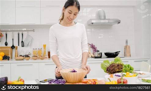 Young Asian woman making salad healthy food in the kitchen, beautiful female in casual use organic vegetables lots of nutrition preparing salad for fit body at home. Healthy food concept.