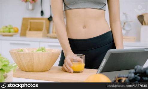 Young Asian woman making salad healthy food while using tablet for looking recipe and drinking orange juice in the kitchen, beautiful female preparing salad for fit body at home. Healthy food concept.
