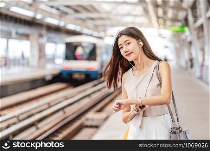 Young Asian woman passenger listening music via smart mobile phone and looking train with Hand watch in subway train station, japanese,chinese,Korean lifestyle, commuter and transportation concept