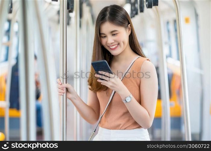 Young Asian woman passenger using smart mobile phone and looking to outside of subway train when traveling in big city, commuter and transportation concept