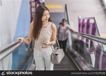 Young Asian woman passenger using smart mobile phone and walking up escalator in subway station when traveling in big city,japanese,chinese,Korean lifestyle and daily life,commuter and transportation