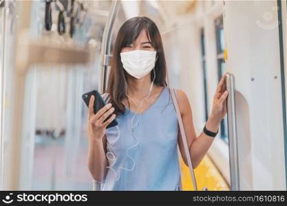 Young Asian woman passenger wearing surgical mask and listening music via smart mobile phone in subway train when traveling in big city at Covid19 outbreak, Infection and Pandemic concept 